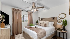 Une chambre avec un lit en bois, un ventilateur de plafond, une télévision, une table de chevet avec une lampe et une fenêtre avec des rideaux.