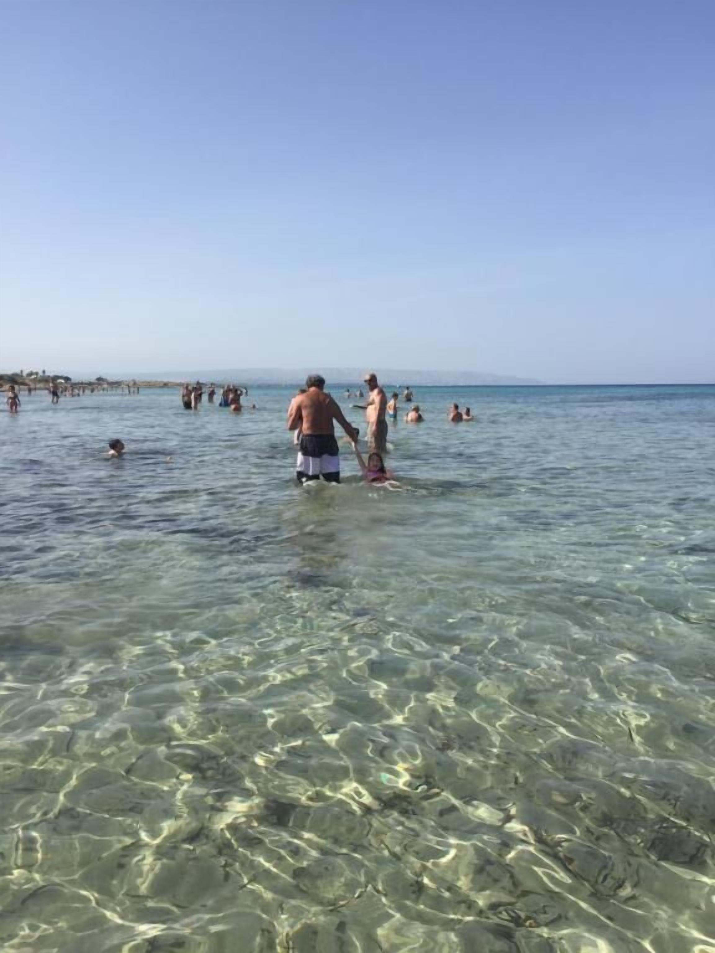 Une plage avec une eau claire et peu profonde, des gens qui nagent et qui se promènent, et une côte visible au loin.