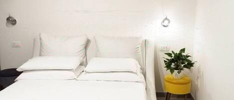 Una cama bien hecha con sábanas y almohadas blancas, una mesita de noche amarilla con una planta, y una pared blanca con textura.