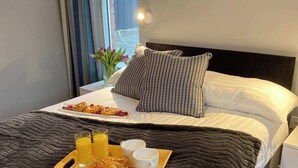 A neatly made bed with a dark quilt, a tray with croissants, fruit, and juice, and a vase with flowers.