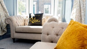 Ein gemütliches Wohnzimmer mit einer Couch und Sesseln, einem 'Relax'-Kissen und Blick auf ein Backsteinhaus durch große Fenster.