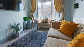 Ein Wohnzimmer mit einer Couch, einem kleinen Beistelltisch mit Pflanze, einem an der Wand montierten Fernseher und einem Fenster mit Vorhängen.