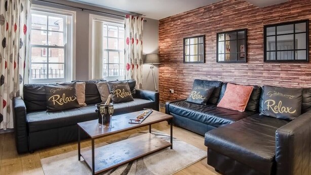 Un salón con dos sofás de cuero negro, cada uno con almohadas que dicen 'Relax', una mesa de centro de madera y un ventanal con cortinas.