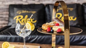 Una bandeja de postres con pasteles y bayas, un vaso de agua con una rodaja de limón, y un sofá negro con almohadas que dicen 'Relax' en un ambiente acogedor.