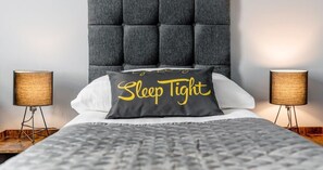 Een bed met een grijs bekleed hoofdeinde, twee bedlampen en een kussen met de tekst 'Sleep Tight'.