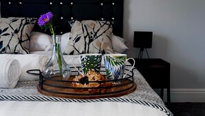 Una cama bien hecha con cabecera negra, una bandeja con galletas y un jarrón de flores, y un cuadro enmarcado sobre la cama.