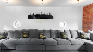 Ein modernes Wohnzimmer mit einem grauen Ecksofa, zwei runden Fenstern, einer städtischen Skyline im Hintergrund und einer Ziegelwand als Blickfang.