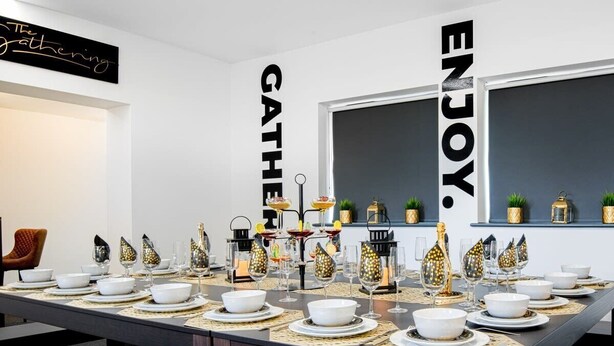 Ein Essbereich mit einem langen Tisch, der für ein Treffen gedeckt ist, mit einem schwarzen Schild an der Wand, auf dem 'The Gathering' und 'ENJOY' stehen.