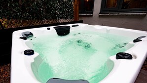 Ein Whirlpool mit integriertem Sitzplatz und Bedienelementen, umgeben von einer Holzterrasse und einem Hecken.