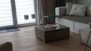A living room with a sofa, a coffee table, and a chair. There is a sliding glass door with blinds, a small table, and a power outlet on the wall. - Modern-gemütlich Eingerichtetes Appartement in Kołobrzeg City (Kolberg)