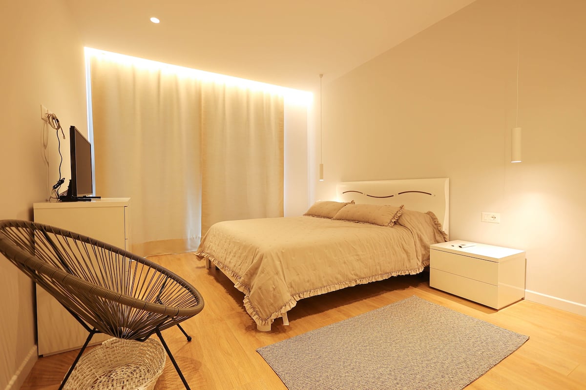 A bedroom with a bed, bedside table, chair, and a large window with curtains.