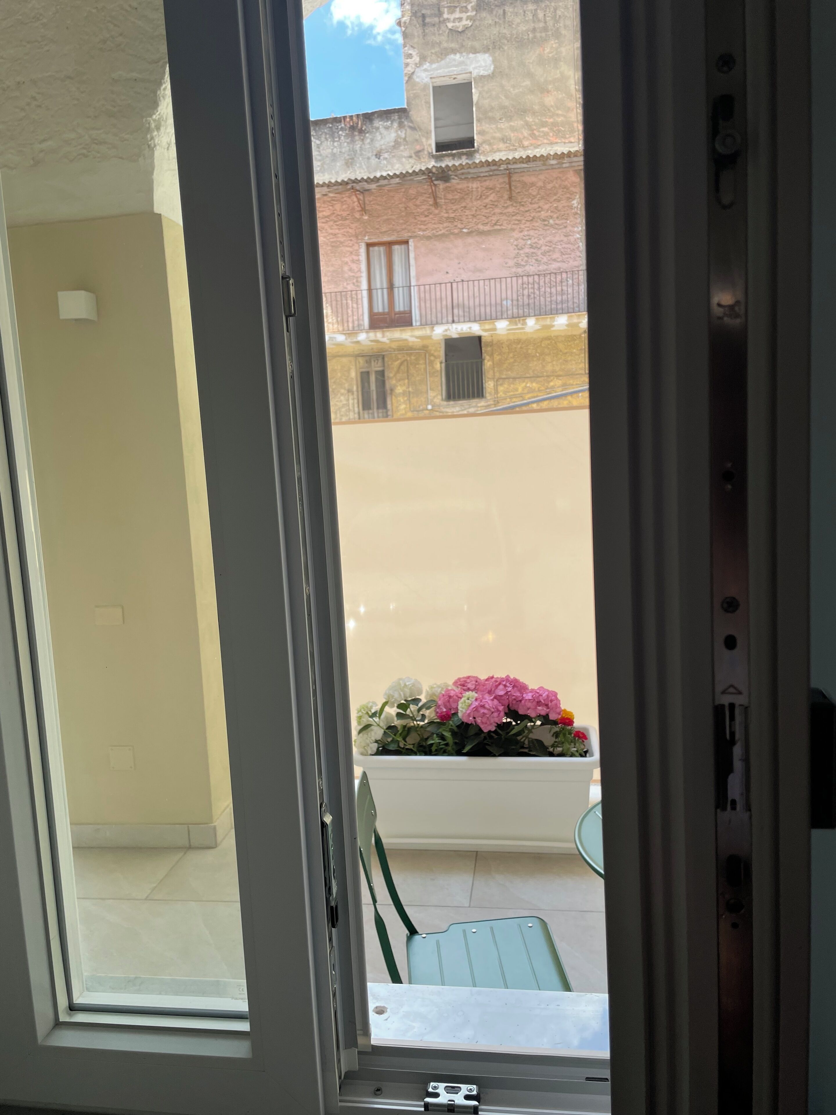 Une vue depuis une fenêtre ornée d’une rambarde en métal, un balcon avec une chaise, et un bâtiment avec un balcon en arrière-plan.