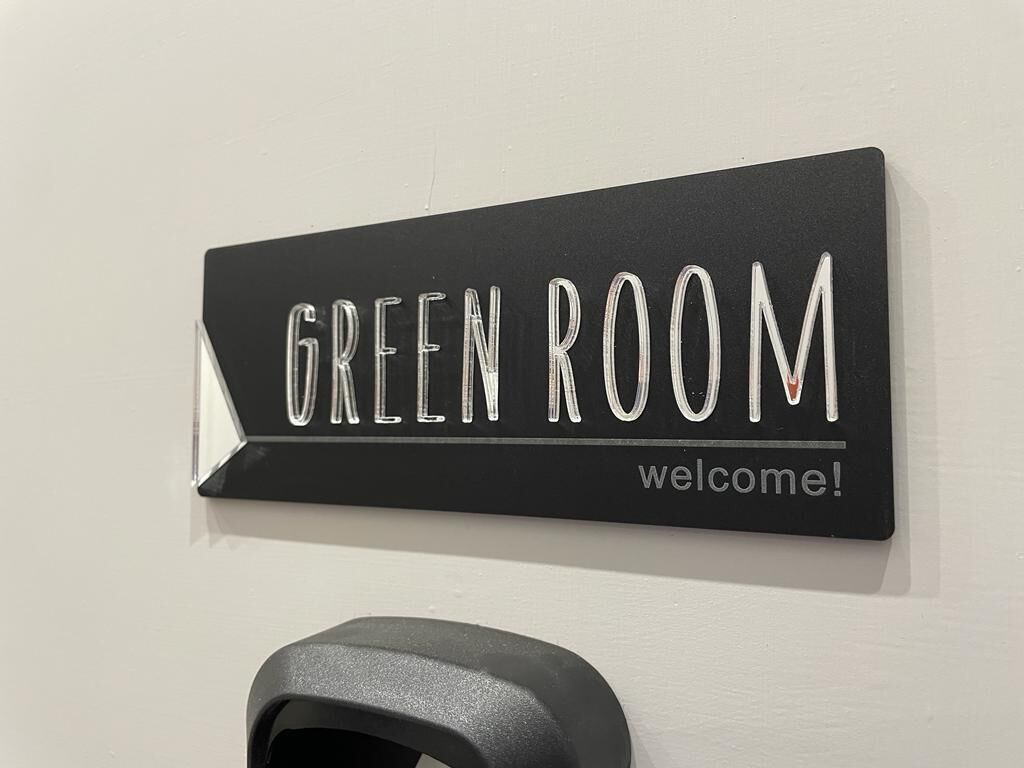 A black sign with white text reading "GREEN ROOM welcome!" mounted on a wall.