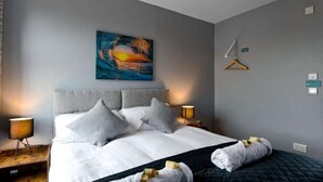 Camera d'albergo con un letto grande, una testiera grigia e un quadro raffigurante un tramonto. C'è un comodino in legno con una lampada e un tavolino anch'esso con una lampada.