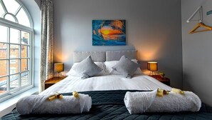 A modern hotel room with a large bed, grey bedding, two bedside lamps, a painting on the wall, and a window with a view of buildings. - The Maelor 2 Bedroomed Apartment |Central Wrexham (Wrexham)