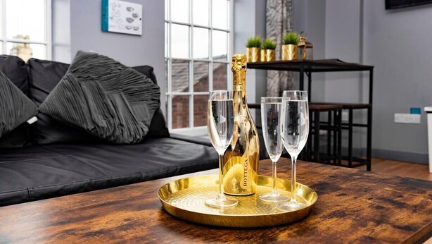 A room with a black leather sofa, a wooden table with a gold tray, a bottle of champagne, and two glasses. - The Maelor 2 Bedroomed Apartment |Central Wrexham (Wrexham)