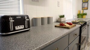A kitchen with a Breville toaster, a granite countertop, a wooden cutting board with tomatoes, and a sink with a plant. - The Flintshire - 3 Bedroomed House - Flint (Flint)