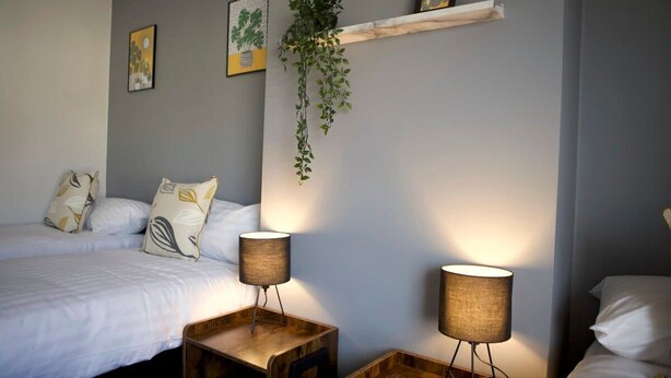 A bedroom with a bed, two bedside tables with lamps, a hanging plant, and a shelf with framed pictures. - The Flintshire - 3 Bedroomed House - Flint (Flint)