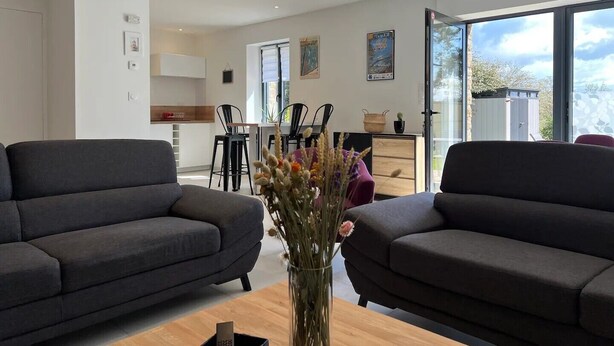 Een moderne woonkamer met twee donkere banken, een houten salontafel en een vaas met gedroogde bloemen. Achtergrond is een eethoek met een tafel en stoelen zichtbaar.