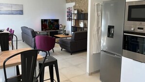 Een moderne woonkamer met een flatscreen televisie, grijze banken, een eethoek met een tafel en stoelen, en een keuken met een roestvrijstalen koelkast.