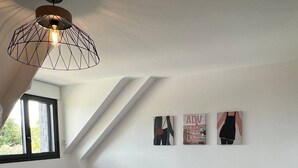 Een slaapkamer met een schuine kap, twee bedden met wit beddengoed, een houten hoofdbord en een geometrische hanglamp. Er zijn drie wanddecoraties en een raam met uitzicht op groen.