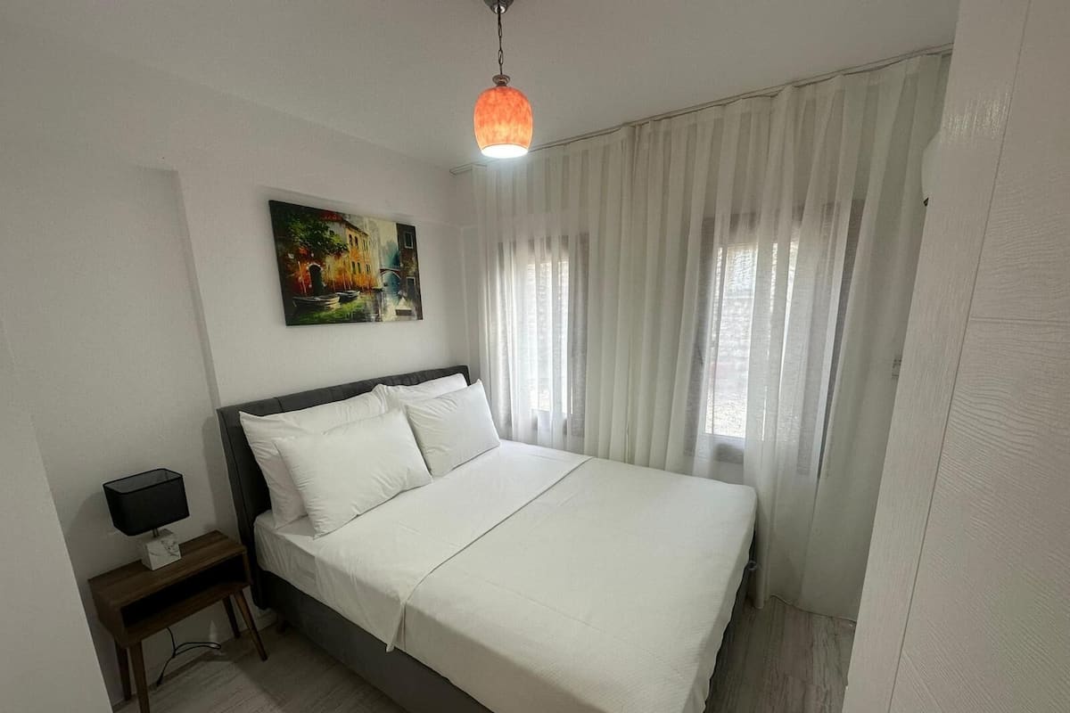 Een netjes ingerichte slaapkamer met een bed, een nachtkastje met een lamp, een schilderij aan de muur en dunne gordijnen voor het raam.