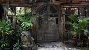 Un bâtiment rustique en bois, doté d’une grande porte en arc de cercle, de plantes en pot et d’une statue en pierre.