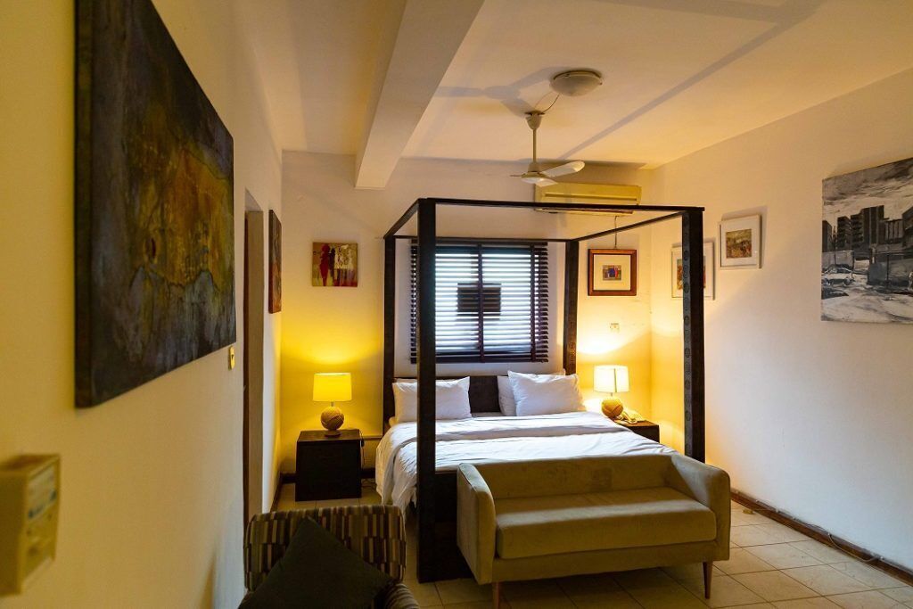 A bedroom with a four-poster bed, bedside lamps, a chair, and artwork on the walls.