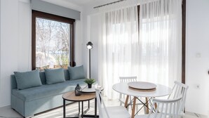 A modern living room with a sofa, a round table, and chairs. There is a window with sheer curtains and a view of trees outside. - Argilos & Argilos Deluxe Residential Complexes-Two - Bedroom Duplex with Garden (Kavala)