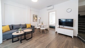 A modern living room with a sofa, coffee table, TV, and a dining area with chairs and a small table. - Argilos & Argilos Deluxe Residential Complexes-Two - Bedroom Duplex with Garden (Kavala)