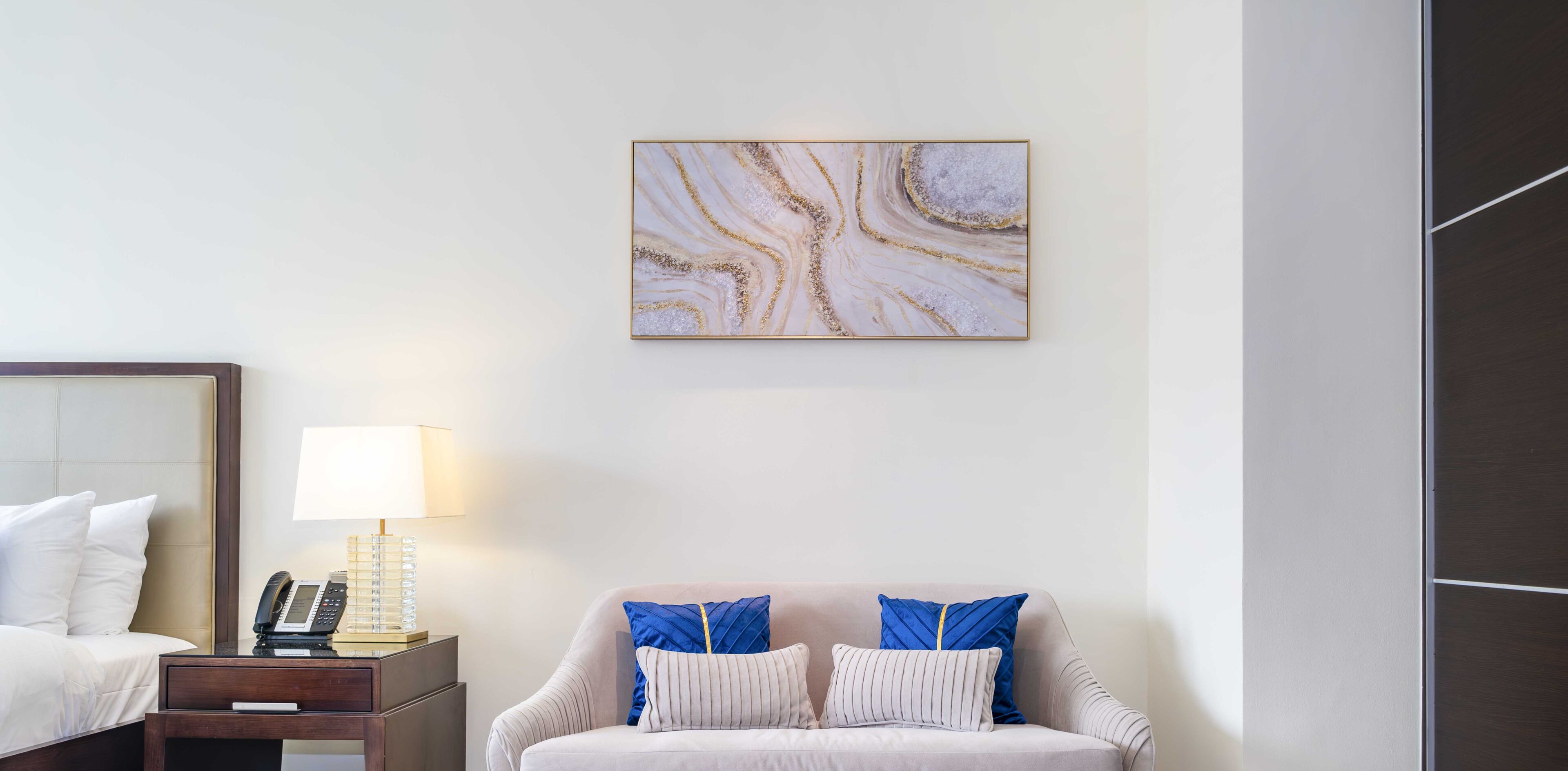 A hotel room with a bed, a bedside table with a lamp, a phone, and a wall-mounted abstract painting.
