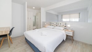 A bedroom with a large bed, a desk, and a chair. There is a window with a blind, a mirror, and a nightstand with a lamp.