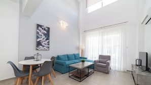 A modern living room with a blue sofa, grey armchair, and a dining table with chairs. There's a painting on the wall and a lamp beside the sofa.