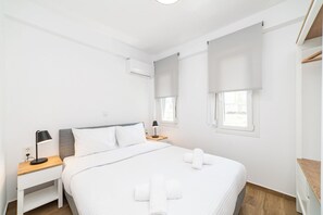 Minimalistinen hotellihuone, jossa on sänky, yöpöydät, työpöytä ja ikkuna, jossa on sälekaihtimet.