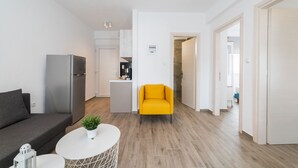 Een moderne woonkamer met een grijze bank, een witte salontafel en een felgele fauteuil. Achtergrond toont een keuken met witte kasten en een roestvrijstalen koelkast.
