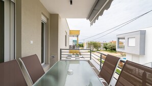 Een balkon met een glazen tafel en stoelen, uitzicht op de omgeving, en een gebouw met een gele luifel.
