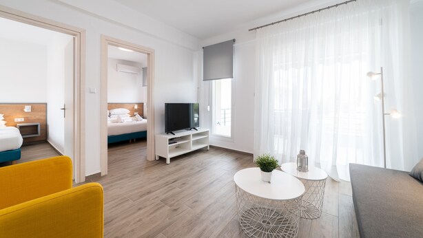 Een moderne hotelkamer met een houten vloer, een bed met televisie, een sofa, een eettafel met stoelen en een groot raam met dunne gordijnen.