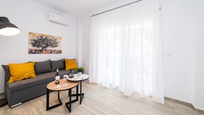 Un salon moderne avec un canapé gris, des coussins jaunes, une table basse ronde sur laquelle se trouvent une bouteille et des verres, et un tableau accroché au mur.