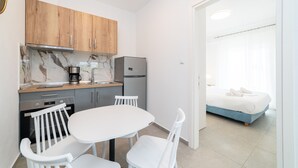 A compact kitchen with wooden cabinets, a marble backsplash, and a refrigerator. There is a small dining table with white chairs. A bed with a blue headboard is visible in the adjacent room. - Nefeli Residential Complex-One Bedroom Apartment (Kavala)