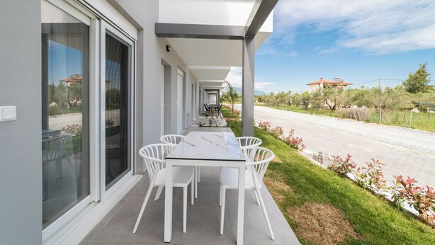 A modern outdoor patio with a table and chairs, a covered walkway, and a view of a road and distant buildings. - Nefeli Residential Complex-One Bedroom Apartment (Kavala)