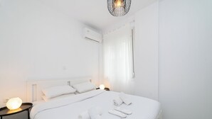 A minimalist hotel room with a large bed, a bedside table with a lamp, and a modern chandelier. - Nefeli Residential Complex-One Bedroom Apartment (Kavala)