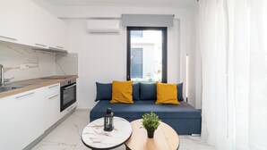 A compact living space with a kitchenette, a sofa with yellow cushions, a small round table, and a potted plant. - Nefeli Residential Complex-One Bedroom Apartment (Kavala)