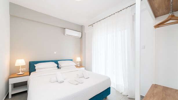 A modern hotel room with a large bed, bedside lamps, a wooden headboard, and a view of the outdoors through sheer curtains. - Nefeli -Two Bedroom Apartment (Kavala)