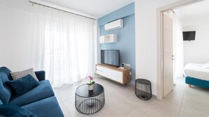 A modern living room with a blue sofa, a small round table with a flower vase, a TV mounted on the wall, and a bed in the adjacent room. - Nefeli -Two Bedroom Apartment (Kavala)