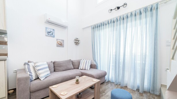 Un salon moderne avec un canapé gris, une table basse en bois et un pouf bleu. Il y a une fenêtre avec des rideaux légers et un climatiseur fixé au mur.