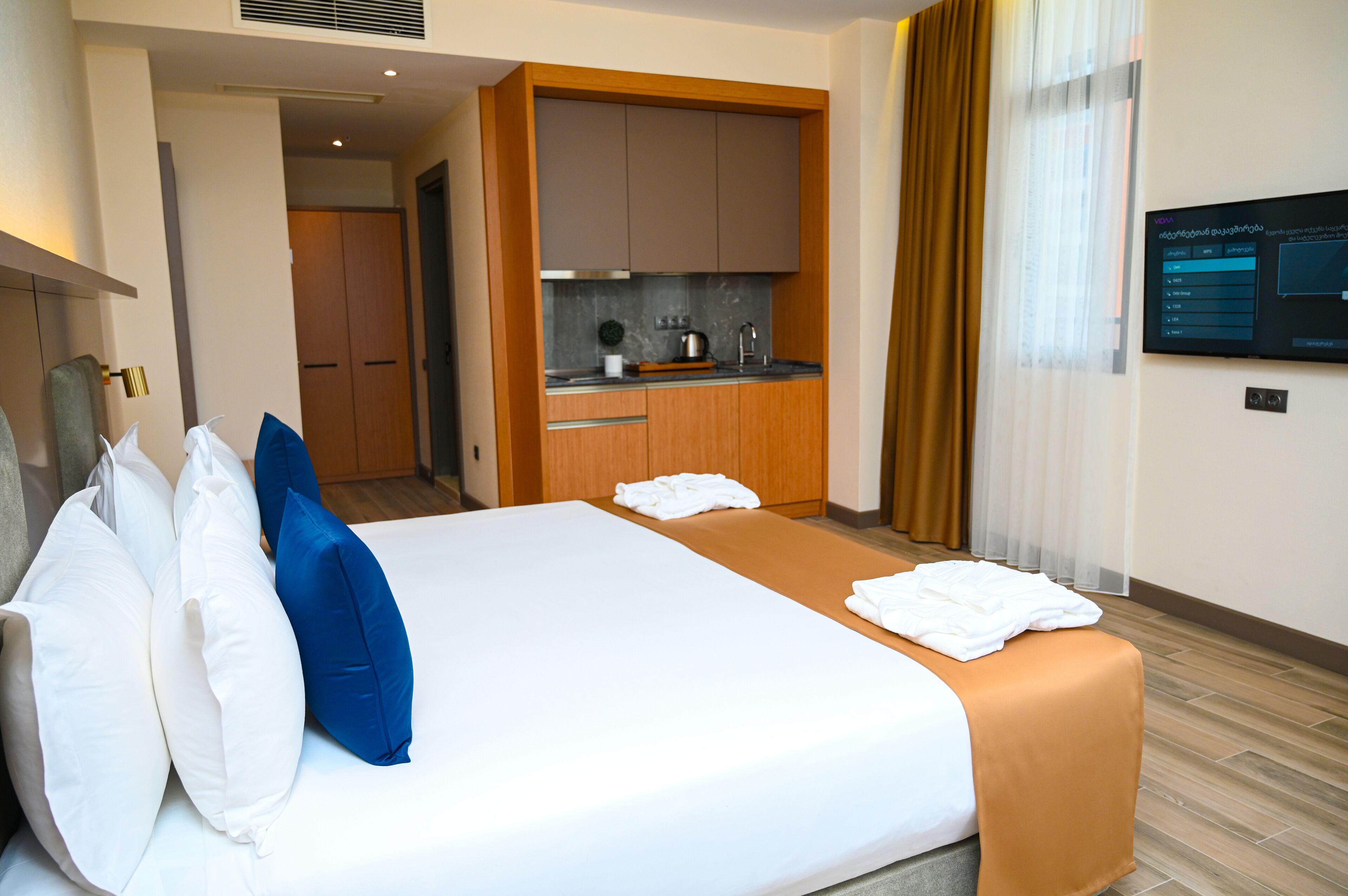 A hotel room with a bed, pillows, a small kitchenette, and a TV on the wall.