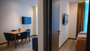 A hotel room with a wooden floor, a bed with a brown cover, a TV mounted on the wall, and a small table with chairs. - New wave apart hotel (Batumi)