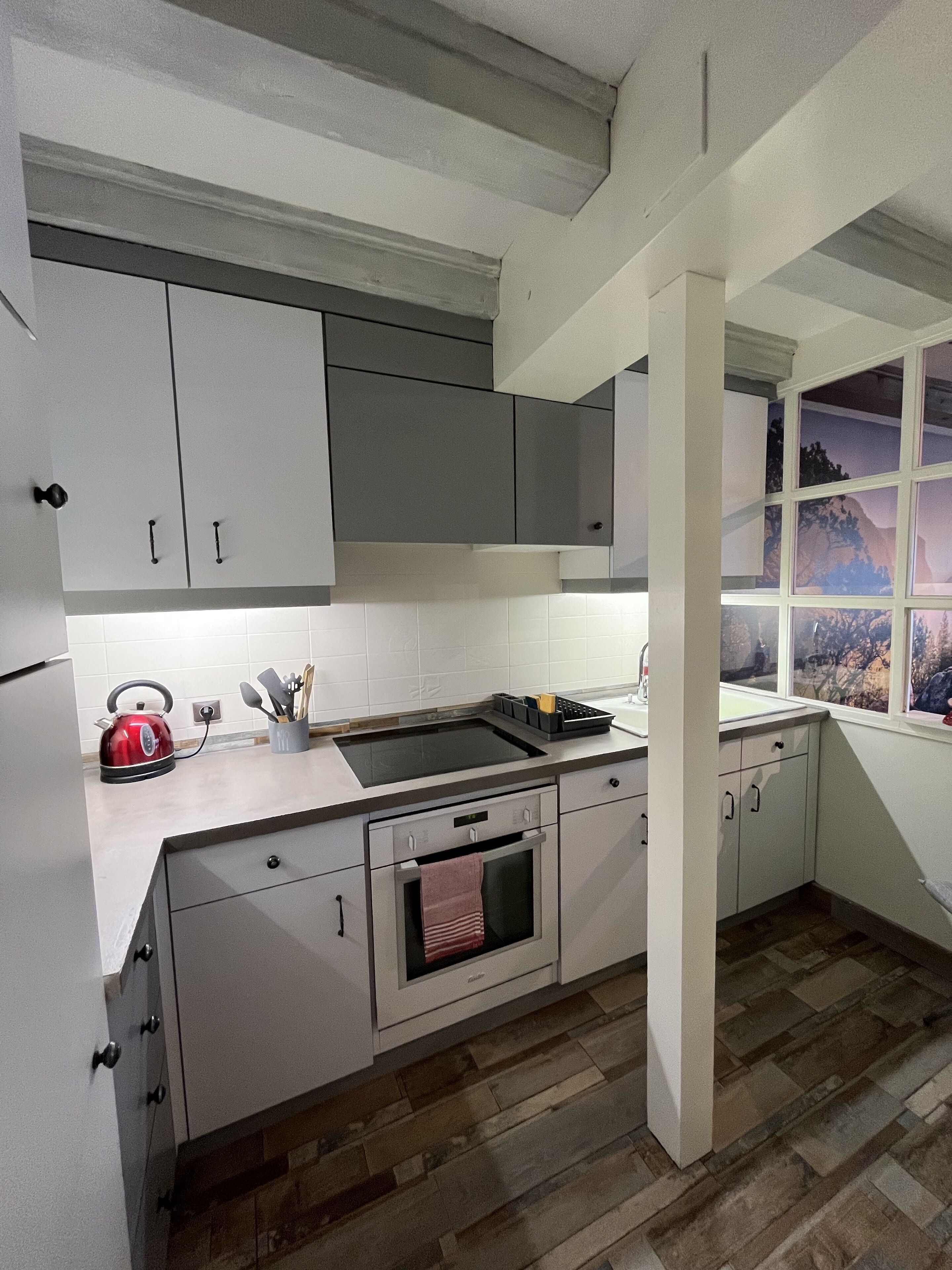 Una cocina con armarios blancos, un hervidor rojo, un horno empotrado y una ventana con vistas a árboles.