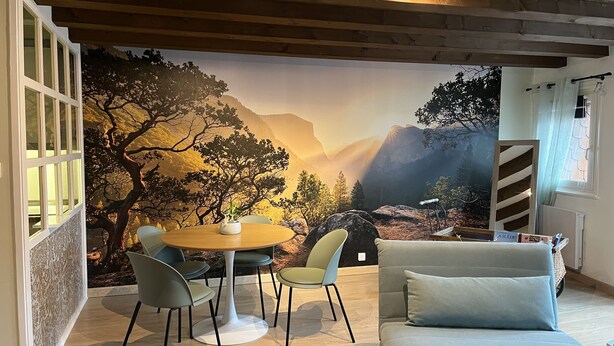 Um quarto acolhedor com teto de madeira, uma mesa de jantar com cadeiras, um sofá e um grande mural de uma paisagem montanhosa.