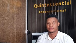 A man seated at a desk with a Panasonic phone and a computer monitor. The background features a dark wooden wall with the logo and name of Quintessential International. - Quintessential Hotel Port Harcourt (Port Harcourt)